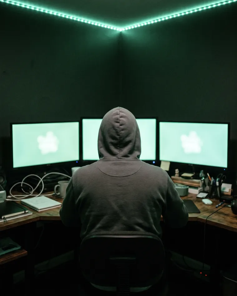 A hooded founder seen from behind at a desk of glowing monitors, face unseen